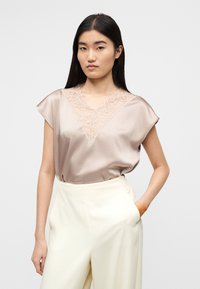 Μπεζ σατέν τοπ με δαντέλα σε σχήμα V στο ντεκολτέ, κοντά μανίκια και χαλαρή εφαρμογή. Συνδυασμένο με κρεμ παντελόνι ψηλής μέσης, με πλαϊνές τσέπες.