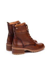 Bottines en cuir marron avec un bout arrondi, des coutures décoratives et des œillets dorés. Elles présentent un petit talon empilé et une semelle texturée.