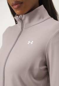 Ljuslila träningsjacka med hög krage, mjukt tyg, hel dragkedja och vit logo på bröstet.