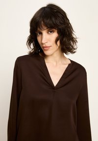 Frau mit welligem dunklem Haar, die ein dunkelbraunes V-Ausschnitt Langarmoberteil trägt und leicht zur Seite schaut, vor einem schlichten hellen Hintergrund.