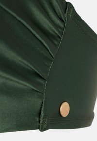 Copricapo in tessuto verde scuro con design arricciato, caratterizzato da un dettaglio in metallo color oro. Texture liscia con bordi cuciti.