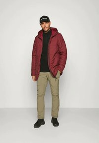 Giubbotto imbottito bordeaux con cappuccio, chiusura con zip, indossato sopra una maglia nera. Abbinato a pantaloni cargo beige leggeri e scarpe nere.