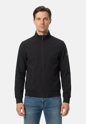 Uomo che indossa una giacca nera con zip e collo alto e jeans azzurri chiari, in piedi davanti a uno sfondo bianco uniforme.