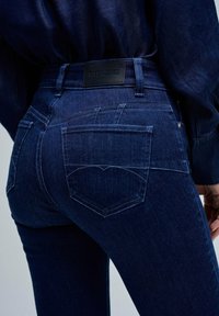 Jeans de denim azul oscuro con una textura lisa, cuentan con dos bolsillos traseros, costuras visibles y una etiqueta negra con "Salsa Jeans" en la cinturilla.