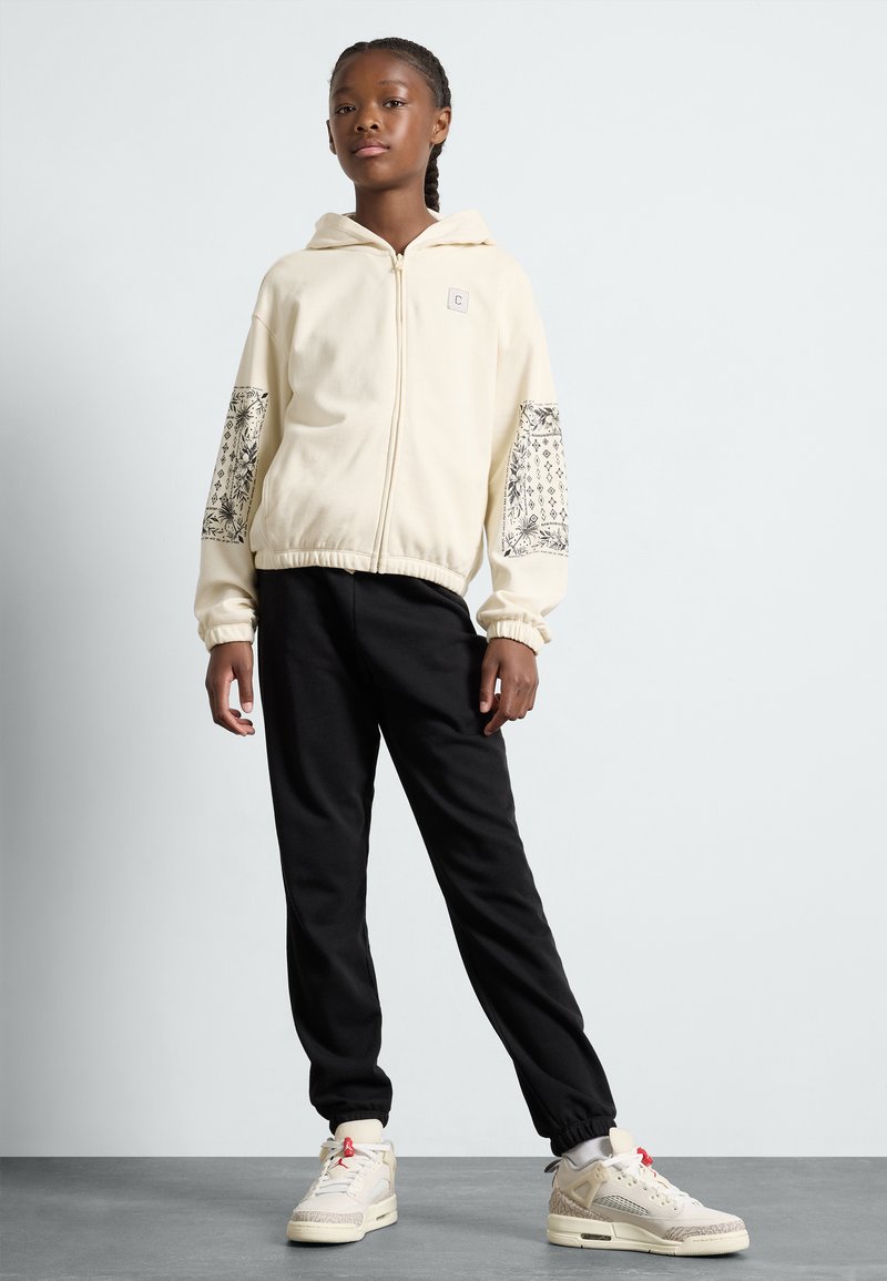 Felpa cream con cerniera e maniche decorative a motivo nero, abbinata a pantaloni jogger neri e sneaker alte di colore chiaro.