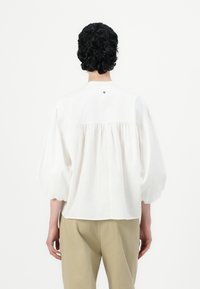 Blusa bianca con maniche a sbuffo, dettagli arricciati sul retro e un piccolo bottone decorativo vicino al collo. Tessuto liscio e leggero.