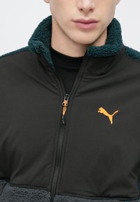 Musta takki, jossa on sinivihreä kaulus ja fleece-kangas. Vetoketjutasku ja oranssi Puma-logo rinnassa. Sileä pinta ja moderni muotoilu.