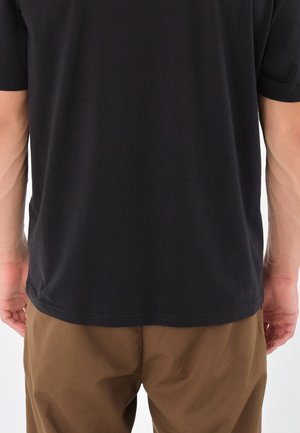 Person, die ein schlichtes schwarzes kurzärmeliges Shirt und braune Hosen trägt, von hinten gezeigt, stehend vor einem weißen Hintergrund.