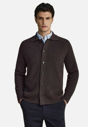 Mand iført en mørkebrun ribbet cardigan med knapper over en lyseblå skjorte med krave med hænderne i lommerne på mørke bukser.