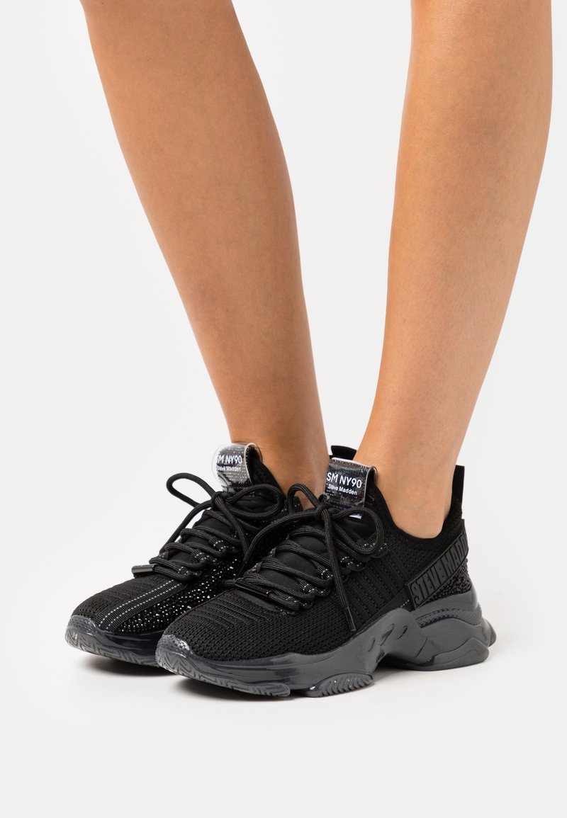 Zapatillas deportivas negras hechas de malla y materiales sintéticos, con un diseño texturizado, cierre con cordones y suelas de goma. Acentos texturizados en los laterales.