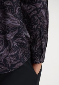Langarmhemd aus dunklem Stoff mit lila botanischem Druck, ausgestattet mit einem knöpfbaren Bündchen und einem geschwungenen Saum. Texturierte, glatte Oberfläche.