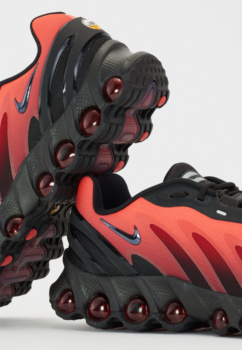 Zapatillas Nike con una parte superior de malla roja y negra, suela de goma negra con cápsulas de aire visibles y acentos metálicos en el logo swoosh.