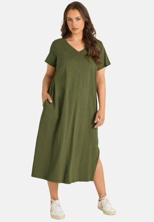 Femme portant une robe midi olive à col en V, manches courtes et poches latérales, associée à des baskets blanches, debout sur un fond uni.