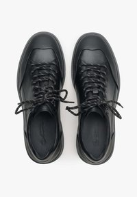 Černé kožené tenisky mají texturovaný povrch, kulatou špičku a ploché tkaničky. Podešev má hladký povrch s minimálním vzorem pro flexibilitu.