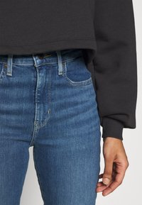 Grå sweatshirt med långa ärmar, kombinerad med högmidjade blå jeans. Jeansarna har en klassisk 5-fickorsdesign och dragkedja fram.