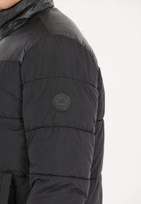 Sort puffjakke med en glat overflade, quiltet tekstur, ribbede mansetter og et cirkulært logo-patch på venstre ærme.