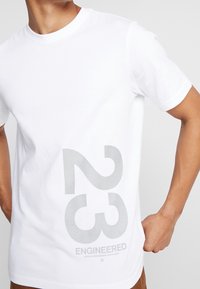 Camiseta de algodón blanca con un gran gráfico gris "23" y la impresión de "ENGINEERED" en la parte frontal. Cuello redondo y mangas cortas.