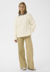 Pull en maille torsadée crème avec un motif texturé et des poignets côtelés, associé à un pantalon large beige et des chaussures de couleur claire.