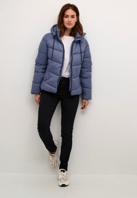 Blue puffer jacket with a hood, quilted design, front zipper, and black drawstrings, paired with black jeans and white sneakers.