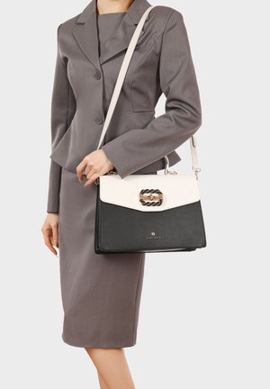 Donna in abito da lavoro grigio che tiene una borsa bianca e nera con chiusura a corda intrecciata dorata e manico superiore sulla spalla.