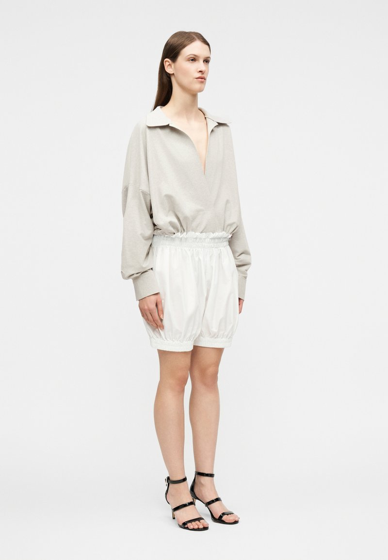 Femme debout portant un haut beige à manches longues avec un col en V profond et un short blanc ample arrivant au genou, ainsi que des chaussures à talons noirs à brides.