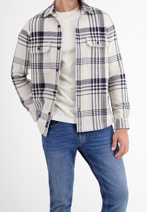 Homme portant une chemise à carreaux blanche et bleu marine sur un t-shirt blanc, avec un jean bleu, debout devant un fond uni.
