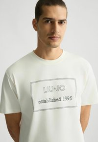 T-shirt bianco in cotone con un logo rettangolare contornato di nero che recita "LIU·JO established 1995" sul davanti. Maniche corte, scollatura rotonda.