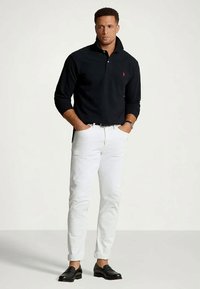 Man står med händerna i fickorna, klädd i en svart långärmad polo, vita uppvikta byxor, svart bälte, klocka och svarta loafers.