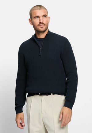 Uomo con capelli corti e barba che indossa un maglione blu navy con mezza zip e pantaloni beige, che guarda leggermente in alto a destra.