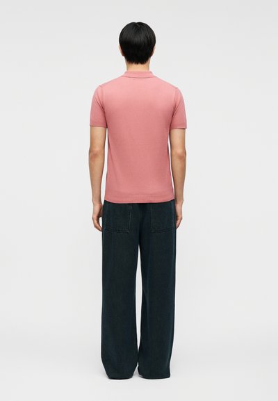 Άτομο που στέκεται με την πλάτη, φοράει κοντομάνικο ροζ πόλο και φαρδιά σκούρα παντελόνια μπροστά σε απλό φόντο.