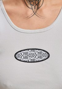 Grijze geribbelde tanktop met een zwart ovale patch met herhalende tekst "BROWEAR" in het wit. Soepele textuur en ronde halslijn.