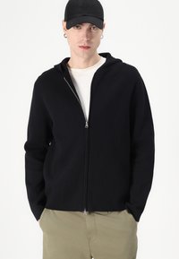 Svart zip-up hoodie med huva, tillverkad av ett mjukt tyg. Har en silverfärgad dragkedja och är kombinerad med en vit crew-neck t-shirt och khakibyxor.