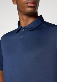 Mørkeblå polo-skjorte laget av teksturert stoff, med krage, tre knapper, og en diskret svart logo på brystet.