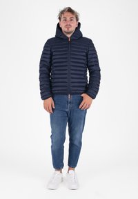 Marineblauwe gewatteerde jas met horizontale naden, capuchon, ritssluiting en oranje ritssluiting. Ggedragen met lichtblauwe jeans en witte sneakers.