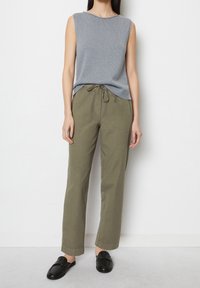 Grijze mouwloze trui met geribbelde textuur, gecombineerd met olijfgroene broek met trekkoord en zwarte instappers. Eenvoudige en casual outfit.