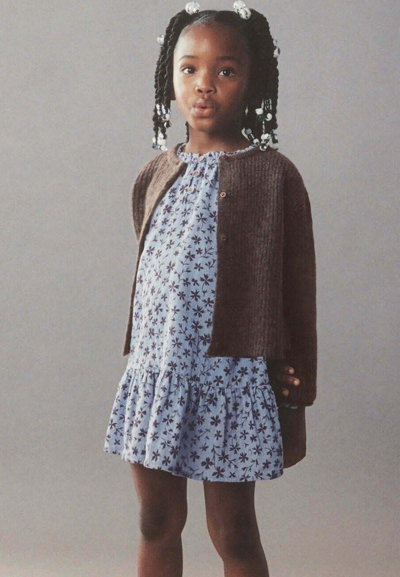 Jeune fille aux cheveux tressés portant un cardigan marron sur une robe fleurie bleu clair, se tient devant un fond uni avec une expression neutre.