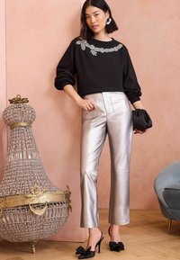 Crna dukserica s ukrašenim izrezom, sjajne srebrne hlače ravnog kroja i crne cipele s potpeticama i mašnama; stoji pored dekorativnog predmeta.