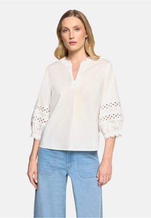 Frau trägt eine weiße Bluse mit Spitzen-Ausschnitten an den aufgebauschten dreiviertellangen Ärmeln und hellblaue, hochtaillierte, weit geschnittene Jeans.