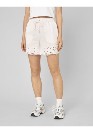 Femme portant un short en dentelle blanche à œillets avec taille à cordon, chaussettes blanches et chaussures de sport blanches, debout devant un fond gris.