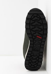 Sort gummi ydersål med et struktureret mønster, grøn øverste materiale og mellemsål med creme-stribet design. Har "TRAXION" branding.