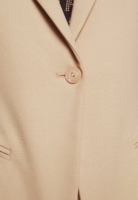 Blazer beige avec une fermeture à un bouton et deux poches latérales. Texture lisse avec un design ajusté, adapté pour des occasions formelles ou décontractées.