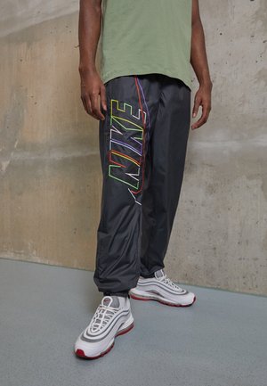 Homme portant un pantalon de survêtement Nike noir avec un logo multicolore et des baskets blanches Nike, debout contre un mur en béton.
