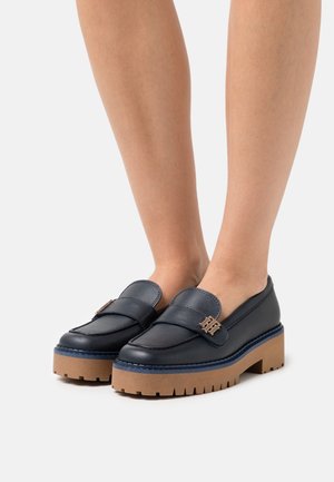 Mocassins en cuir marine avec une plateforme basse, présentant un accent décoratif en métal doré et des coutures contrastantes bleues.