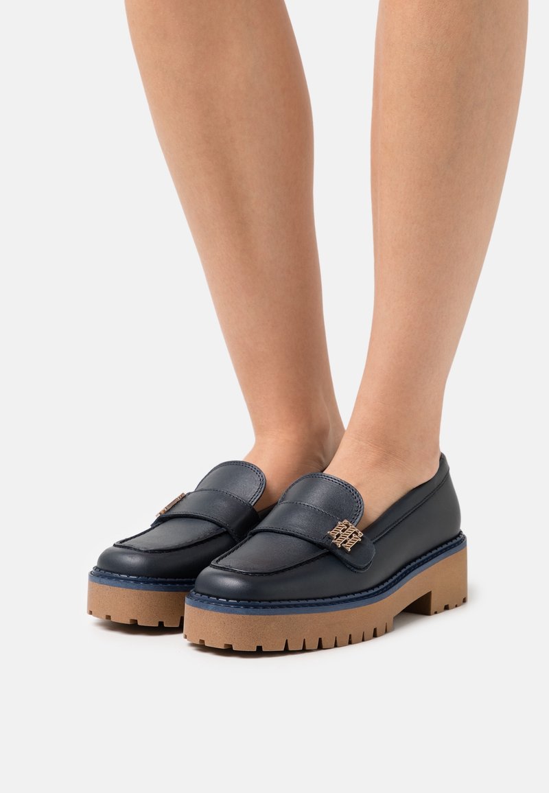 Mocassins en cuir marine avec une plateforme basse, présentant un accent décoratif en métal doré et des coutures contrastantes bleues.