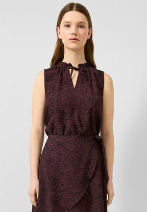 Junge Frau mit glatten braunen Haaren, die ein ärmelloses dunkelviolettes Kleid mit einem schwarzen wirbelnden Muster und einem gebundenen Ausschnitt trägt.