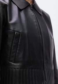Μαύρο δερμάτινο μπουφάν με κλείσιμο με φερμουάρ, μυτερό γιακά και ριμπ στη βάση. Διαθέτει πλαϊνή τσέπη και λεία, ελαφρώς γυαλιστερή υφή.