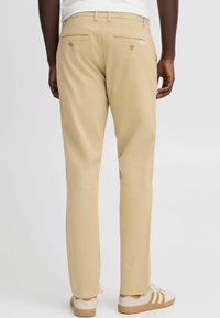 Beige chinos with a straight-leg fit, featuring double back pockets with button closures. Worn with white sneakers with tan accents.