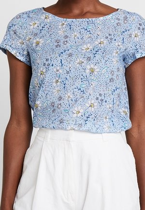 Lichtblauw bloemenblouse met korte mouwen, gecombineerd met hoge witte broek, gedragen door een persoon met een donkere huidskleur.