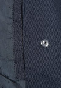 Primer plano de tela azul marino con un acabado texturizado, botón transparente y costuras visibles. La tela muestra un patrón de tejido diagonal.