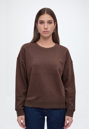 Jonge vrouw met lang bruin haar draagt gouden oorbellen, een bruin sweatshirt en donkerblauwe jeans, staand tegen een effen lichte achtergrond.